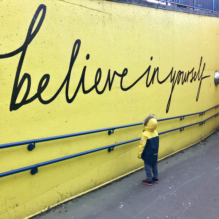 Kind in Regenmantel vor einer Wand mit Überschrift 'Believe in yourself'